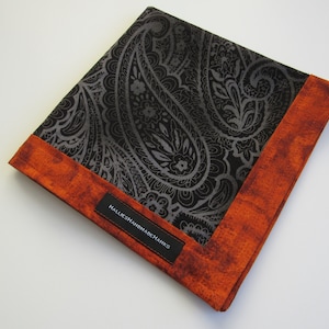 May include: A square pocket square featuring a central black fabric panel with a dark grey paisley pattern. The fabric is bordered by a textured, mottled orange fabric trim. A black rectangular label with the white text HALLIESHANDMADEHANKS is sewn onto the orange border.