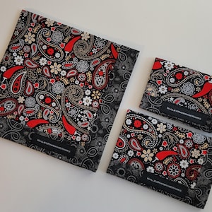 May include: A set of three fabric items with a black, red, and white paisley pattern. The largest item is rectangular, and the other two are smaller rectangles. Each item has a black border with a dotted design and a label that reads "HALLIESHANDMADEHANKS."