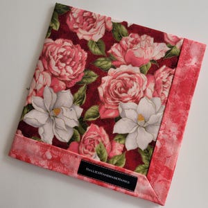 May include: Square floral handkerchief featuring a pattern of pink roses and white magnolias on a burgundy background. The edges are trimmed with a pink floral fabric. A black tag reads "HALLIESHANDMADEHANKS".