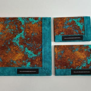 May include: Three fabric squares featuring a mottled rust-brown and turquoise pattern with solid teal borders. Each piece includes a black rectangular label with the white text HALLIESHANDMADEHANKS stitched onto the bottom edge of the border.
