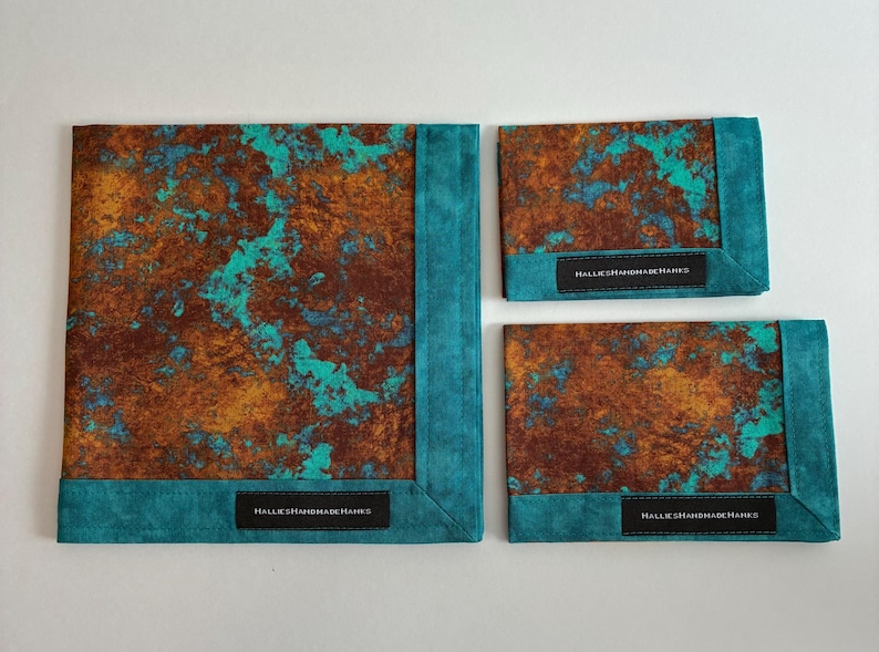 May include: Three fabric squares featuring a mottled rust-brown and turquoise pattern with solid teal borders. Each piece includes a black rectangular label with the white text HALLIESHANDMADEHANKS stitched onto the bottom edge of the border.