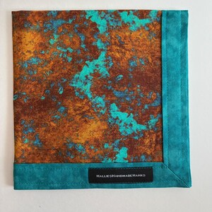 May include: A square fabric handkerchief featuring a mottled pattern of deep brown, copper, and bright turquoise colors. The piece is finished with a solid turquoise border and a black fabric label reading HALLIESHANDMADEHANKS in white text.