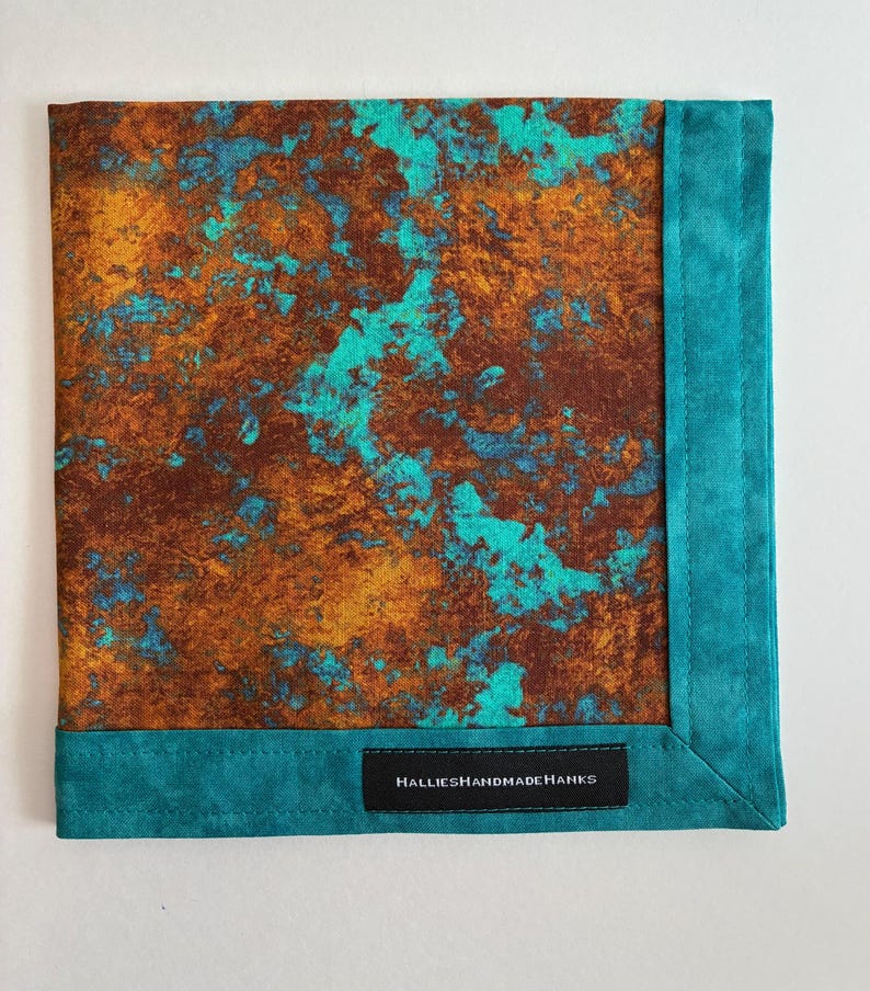 May include: A square fabric handkerchief featuring a mottled pattern of deep brown, copper, and bright turquoise colors. The piece is finished with a solid turquoise border and a black fabric label reading HALLIESHANDMADEHANKS in white text.