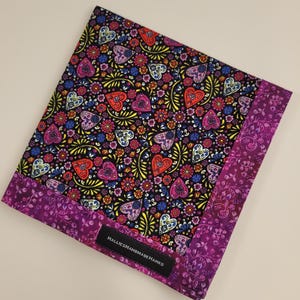 May include: A square, patterned textile with a black background, featuring colorful heart and floral designs. The border is a deep purple with a floral pattern. The label reads "HALLIESHANDMADE HANKS".