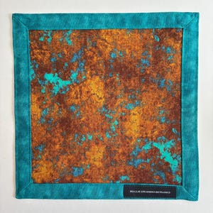 May include: A square fabric handkerchief featuring a mottled, rust-colored pattern with vibrant turquoise accents. The center panel is framed by a solid turquoise border. A small black rectangular tag with the white text HALLIESHANDMADEHANKS is sewn onto the bottom right corner.