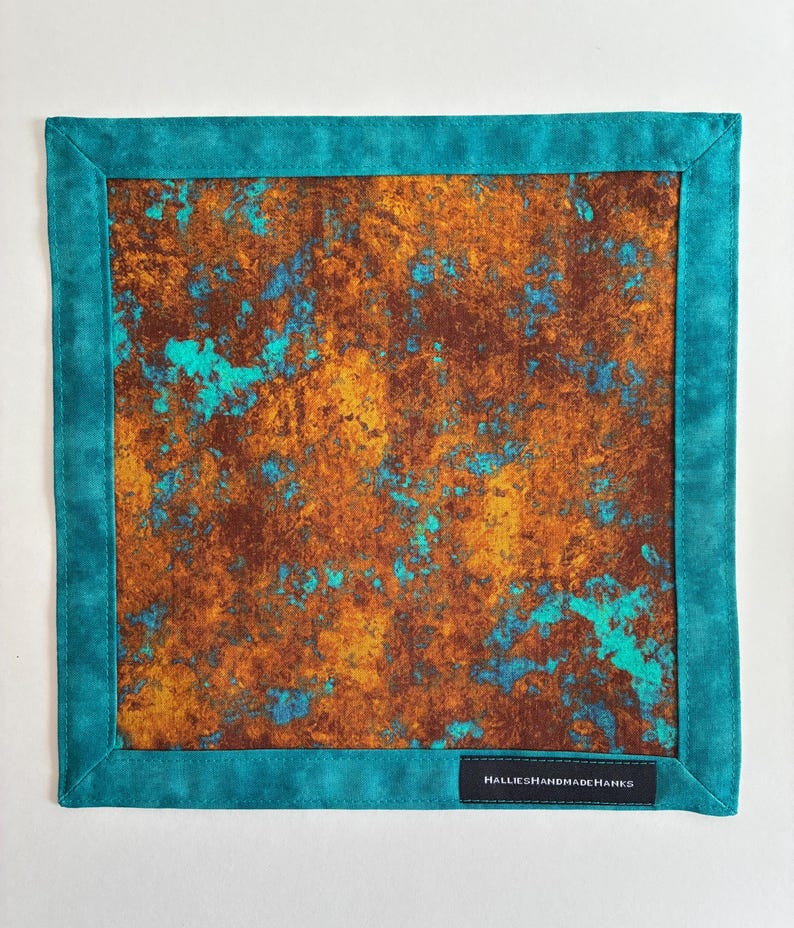 May include: A square fabric handkerchief featuring a mottled, rust-colored pattern with vibrant turquoise accents. The center panel is framed by a solid turquoise border. A small black rectangular tag with the white text HALLIESHANDMADEHANKS is sewn onto the bottom right corner.