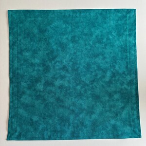 May include: A square fabric napkin in a deep teal color with a mottled, cloud-like pattern. The napkin features a stitched border running parallel to all four edges, creating a framed effect on the surface of the textile material.