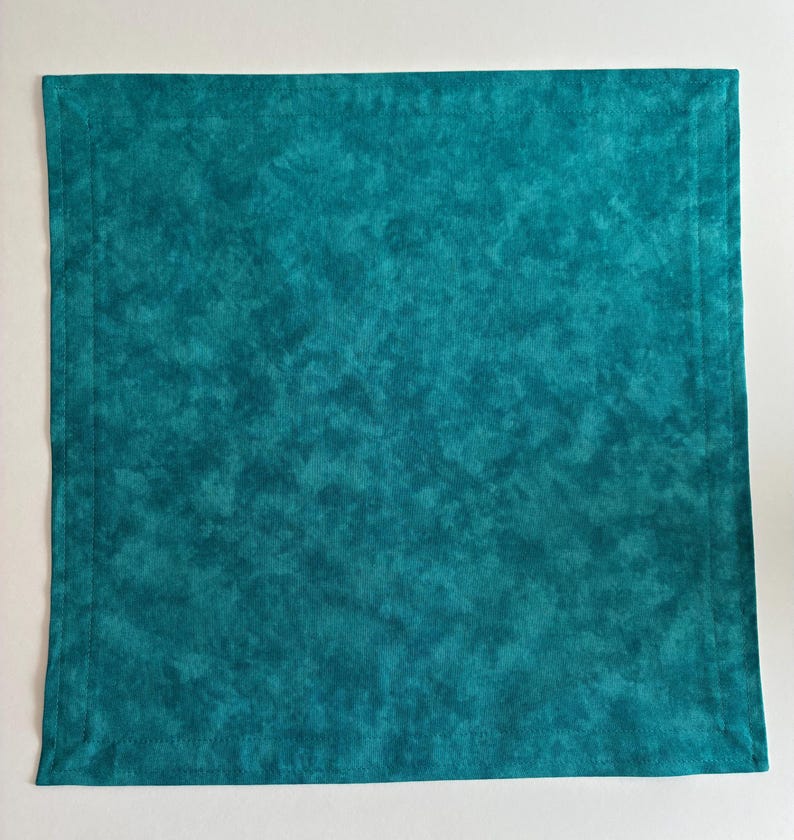 May include: A square fabric napkin in a deep teal color with a mottled, cloud-like pattern. The napkin features a stitched border running parallel to all four edges, creating a framed effect on the surface of the textile material.
