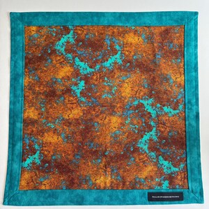 May include: A square fabric cloth featuring a central panel with a mottled, rust-like pattern in shades of deep brown, orange, and bright turquoise. The cloth is framed by a solid teal border and includes a small black label reading HALLIESHANDMADEHANKS.