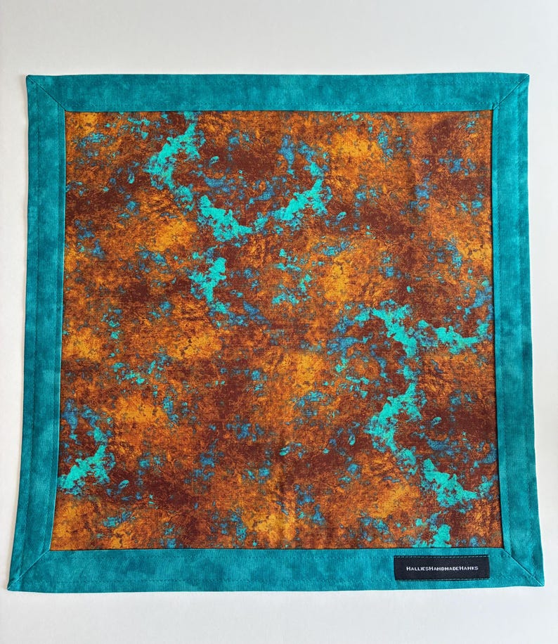 May include: A square fabric cloth featuring a central panel with a mottled, rust-like pattern in shades of deep brown, orange, and bright turquoise. The cloth is framed by a solid teal border and includes a small black label reading HALLIESHANDMADEHANKS.