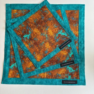 May include: Three square fabric handkerchiefs featuring a mottled rust-brown and bright turquoise pattern. Each piece has a solid teal border and a small black rectangular tag with the white text HALLIESHANDMADEHANKS sewn onto the corner.