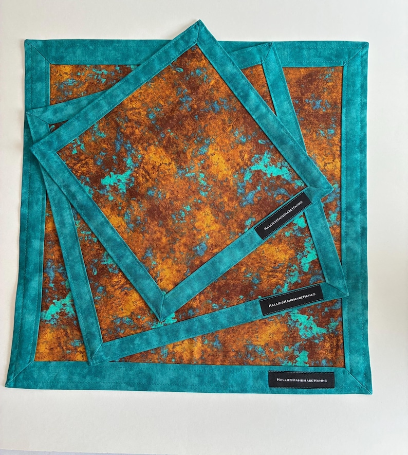 May include: Three square fabric handkerchiefs featuring a mottled rust-brown and bright turquoise pattern. Each piece has a solid teal border and a small black rectangular tag with the white text HALLIESHANDMADEHANKS sewn onto the corner.
