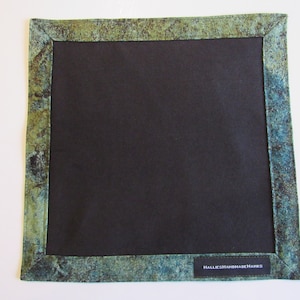 May include: A square black placemat with a green and blue patterned border. The border has a mottled, marbled look. The placemat has a label that reads "Hallie's Handmade Hanes".
