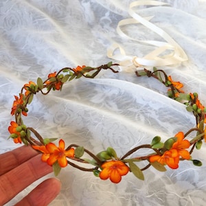May include: A floral crown with orange flowers and green leaves on a brown vine base. The crown is tied with a white ribbon.