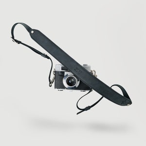 May include: A black leather camera strap with the initials "JPV" embossed on it. The strap is attached to a silver camera.