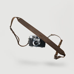 May include: A brown leather camera strap with the initials "JPV" embossed on it. The strap is attached to a silver camera.