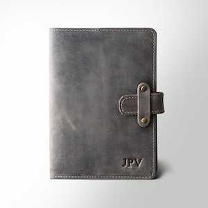 May include: Gray leather journal with a strap closure and brass-colored rivets. The journal has the initials "JPV" embossed on the lower right corner. The leather has a distressed look with visible grain and texture, suggesting a vintage or rustic style.