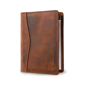 May include: A brown leather notebook cover with a flap and a stitched edge. The cover has a personalized monogram "TRO" embossed on the front.