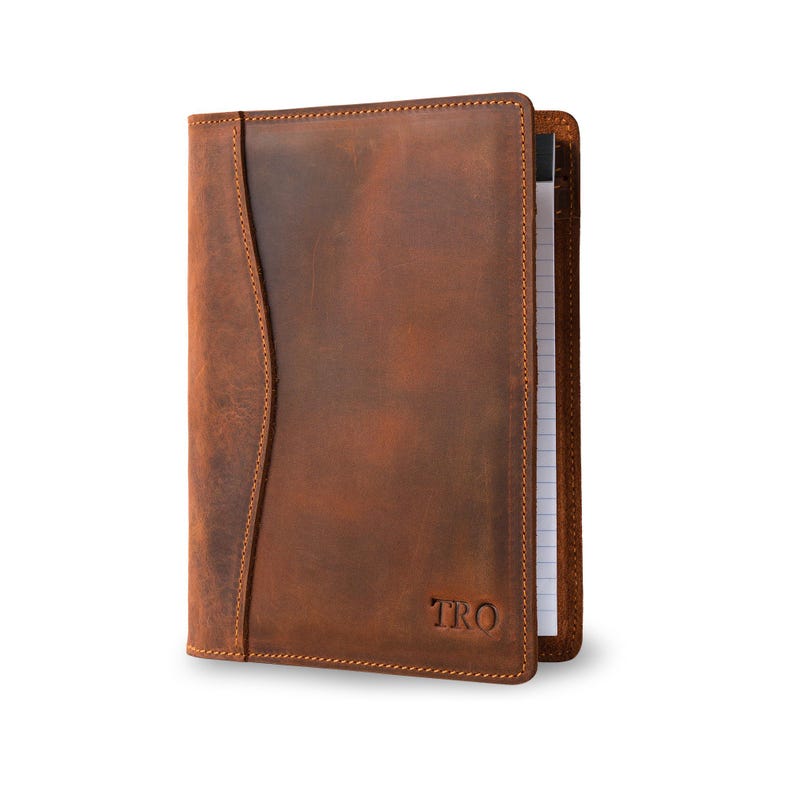 May include: A brown leather notebook cover with a flap and a stitched edge. The cover has a personalized monogram "TRO" embossed on the front.