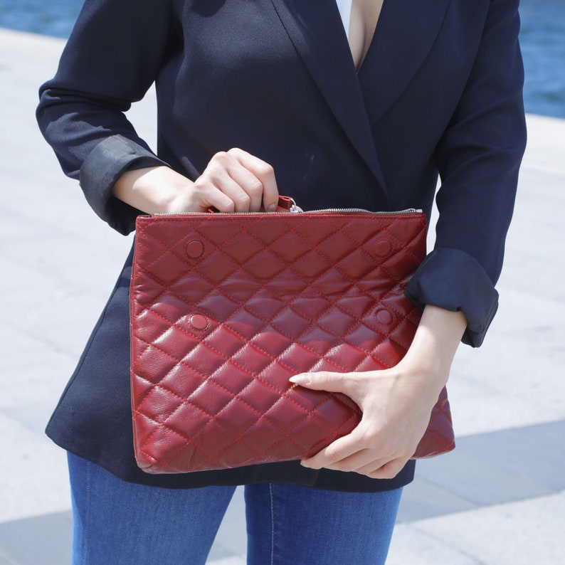 Op de afbeelding: Een rode quilted clutch met een ritssluiting. De tas heeft een ruitpatroon en wordt vastgehouden door iemand die een donkerblauwe blazer draagt.