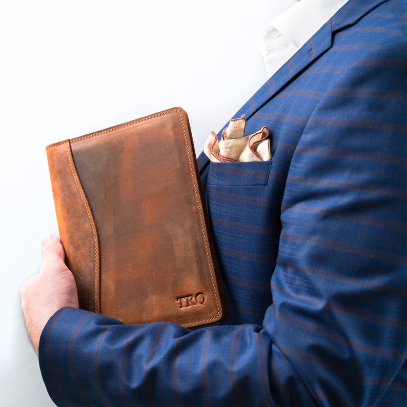 May include: A brown leather notebook with the initials "TRQ" embossed on the front. The notebook is being held by a person wearing a blue blazer.