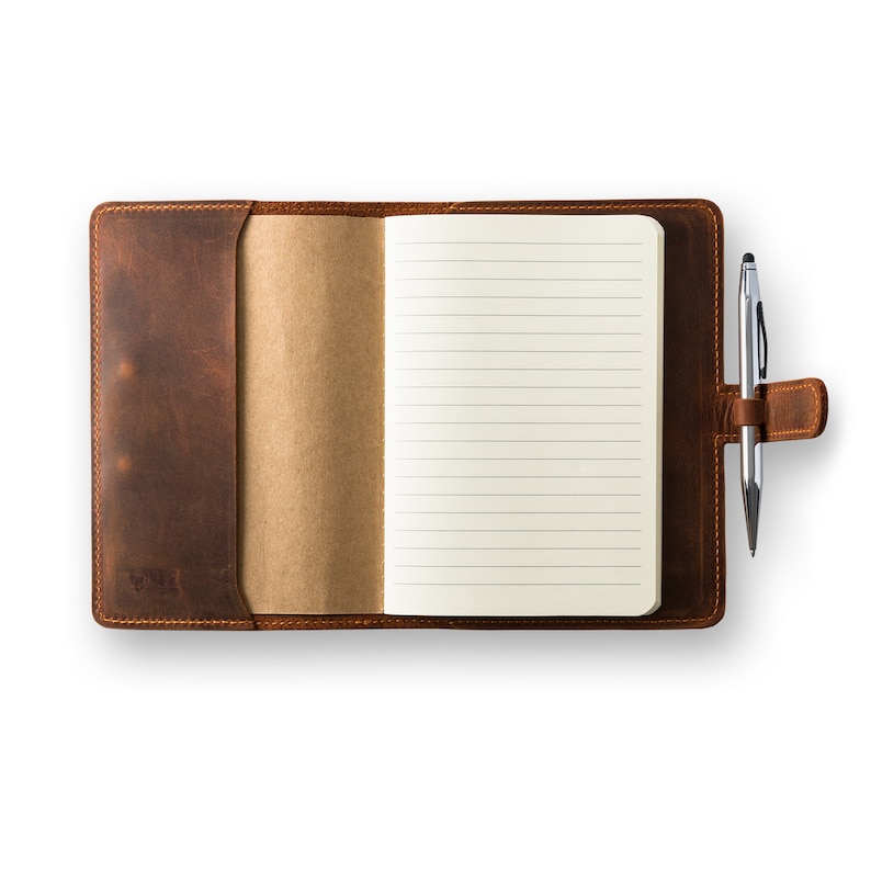Op de afbeelding: Een bruin lederen gebonden dagboek met een pen die in de leren band is gestoken. Het dagboek is open op een blanco pagina met gestreept papier.