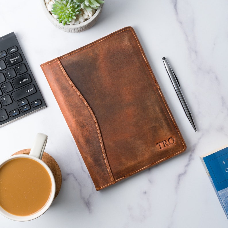 May include: A brown leather journal with a flap closure and a personalized inscription that reads "TRO". The journal is on a white marble surface with a keyboard, a cup of coffee, a pen, and a book.