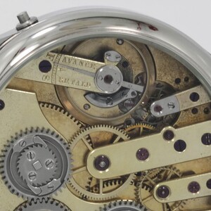 May include: Close-up view of the inner workings of a silver pocket watch with a gold-toned face. The watch has a lever set mechanism and the words "Advance" and "Retard" are visible on the face.