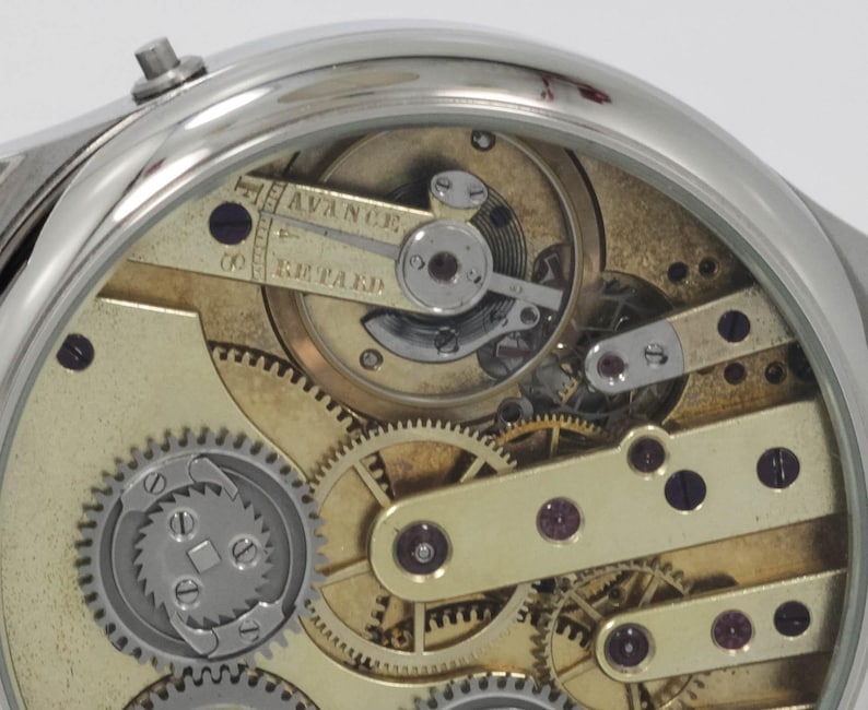 May include: Close-up view of the inner workings of a silver pocket watch with a gold-toned face. The watch has a lever set mechanism and the words "Advance" and "Retard" are visible on the face.