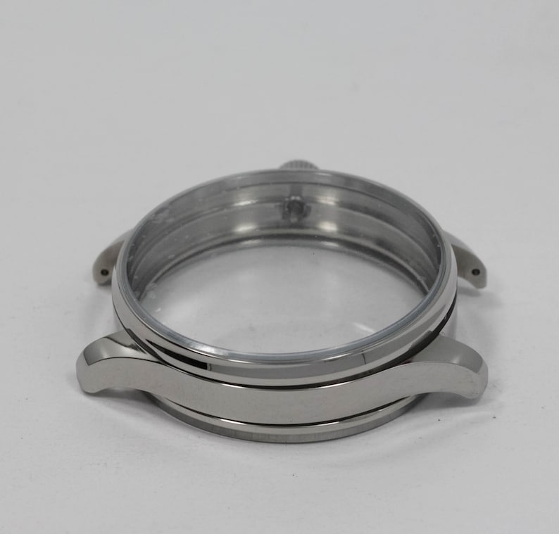 May include: A silver-toned watch case with a clear glass face. The case has a circular design with curved lugs for the watch band. The watch case is made of metal and has a polished finish.