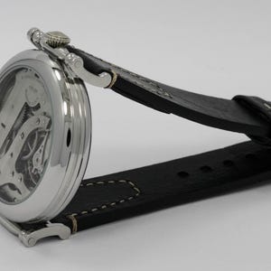 K&ouml;nnte beinhalten: Eine silberfarbene Armbanduhr mit skelettiertem Zifferblatt, das interne mechanische Zahnr&auml;der und Komponenten freilegt. Die Uhr verf&uuml;gt &uuml;ber ein poliertes rundes Geh&auml;use und ein schwarzes Lederarmband mit sichtbaren wei&szlig;en N&auml;hten.