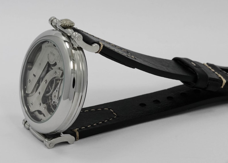 K&ouml;nnte beinhalten: Eine silberfarbene Armbanduhr mit skelettiertem Zifferblatt, das interne mechanische Zahnr&auml;der und Komponenten freilegt. Die Uhr verf&uuml;gt &uuml;ber ein poliertes rundes Geh&auml;use und ein schwarzes Lederarmband mit sichtbaren wei&szlig;en N&auml;hten.