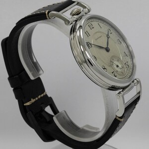 K&ouml;nnte beinhalten: Eine silberfarbene Armbanduhr mit einem runden, cremefarbenen Zifferblatt, schwarzen arabischen Ziffern und dem Schriftzug Howard. Die Uhr hat schwarze Zeiger, ein kleines Hilfszifferblatt auf der Sechs-Uhr-Position und ein schwarzes Lederarmband.