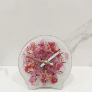 May include: A pink and white glass clock with silver hands and a white base. The clock face is made of broken glass pieces that have been fused together. The clock face has the text "Jellyglass" on it.