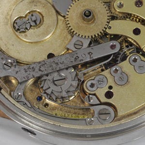 May include: Close-up of a complex watch mechanism with gold-toned gears and a silver-toned lever. The mechanism is intricate and detailed, showcasing the craftsmanship of watchmaking.
