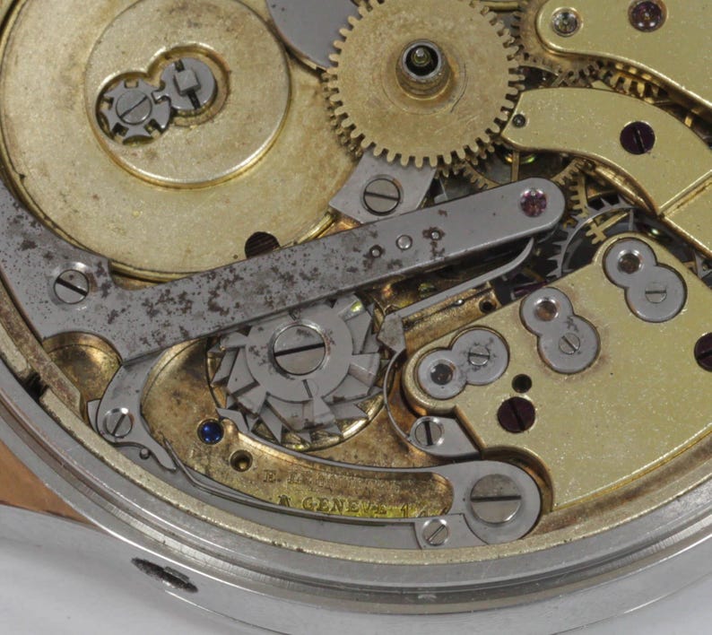 May include: Close-up of a complex watch mechanism with gold-toned gears and a silver-toned lever. The mechanism is intricate and detailed, showcasing the craftsmanship of watchmaking.