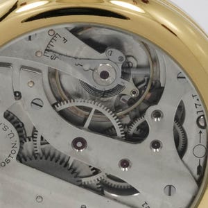 May include: Close-up of a gold-toned pocket watch mechanism. The intricate internal workings are visible, including gears, springs, and other components. The watch face is absent, revealing the detailed mechanical parts. The watch is a vintage item.