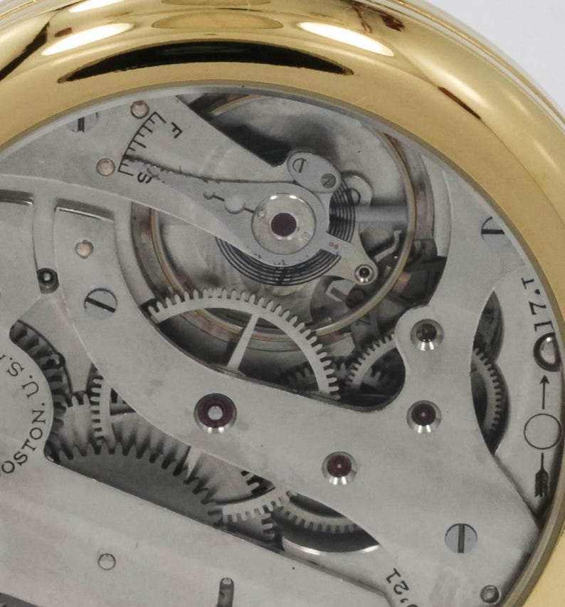 May include: Close-up of a gold-toned pocket watch mechanism. The intricate internal workings are visible, including gears, springs, and other components. The watch face is absent, revealing the detailed mechanical parts. The watch is a vintage item.