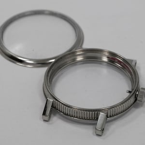 May include: A disassembled watch case with a clear glass crystal and a silver-colored, ridged bezel. The case has two lugs for a watch band and a small crown. The crystal is separate from the case.