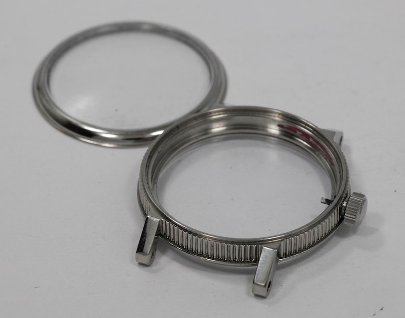 May include: A disassembled watch case with a clear glass crystal and a silver-colored, ridged bezel. The case has two lugs for a watch band and a small crown. The crystal is separate from the case.
