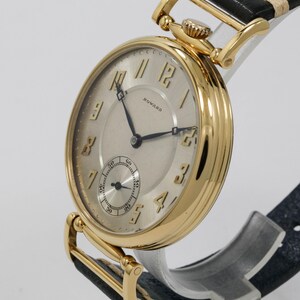 May include: A gold wristwatch with a cream face and black hands. The watch has a subdial and the name "HOWARD" is printed on the face. The watch has a black leather strap with gold-coloured accents.