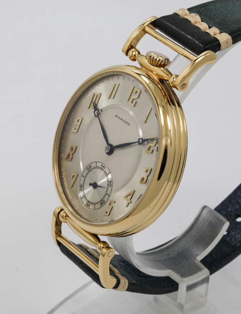 May include: A gold wristwatch with a cream face and black hands. The watch has a subdial and the name "HOWARD" is printed on the face. The watch has a black leather strap with gold-coloured accents.