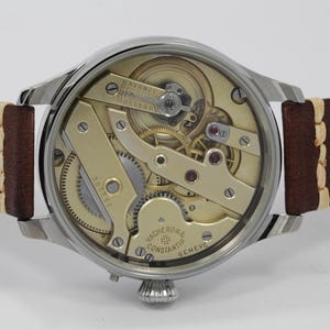 Puede incluir: La parte posterior de un reloj de pulsera plateado con una correa de cuero marr&oacute;n. La esfera del reloj est&aacute; abierta, revelando el intrincado funcionamiento interno del reloj. La esfera del reloj est&aacute; etiquetada como "Vacheron Constantin Geneve".