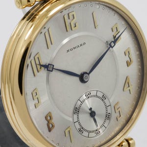 May include: A gold pocket watch with a silver face. The watch has black hands, gold numerals, and a small seconds dial. The word "HOWARD" is printed on the face. The watch has a gold crown and a gold chain attachment.