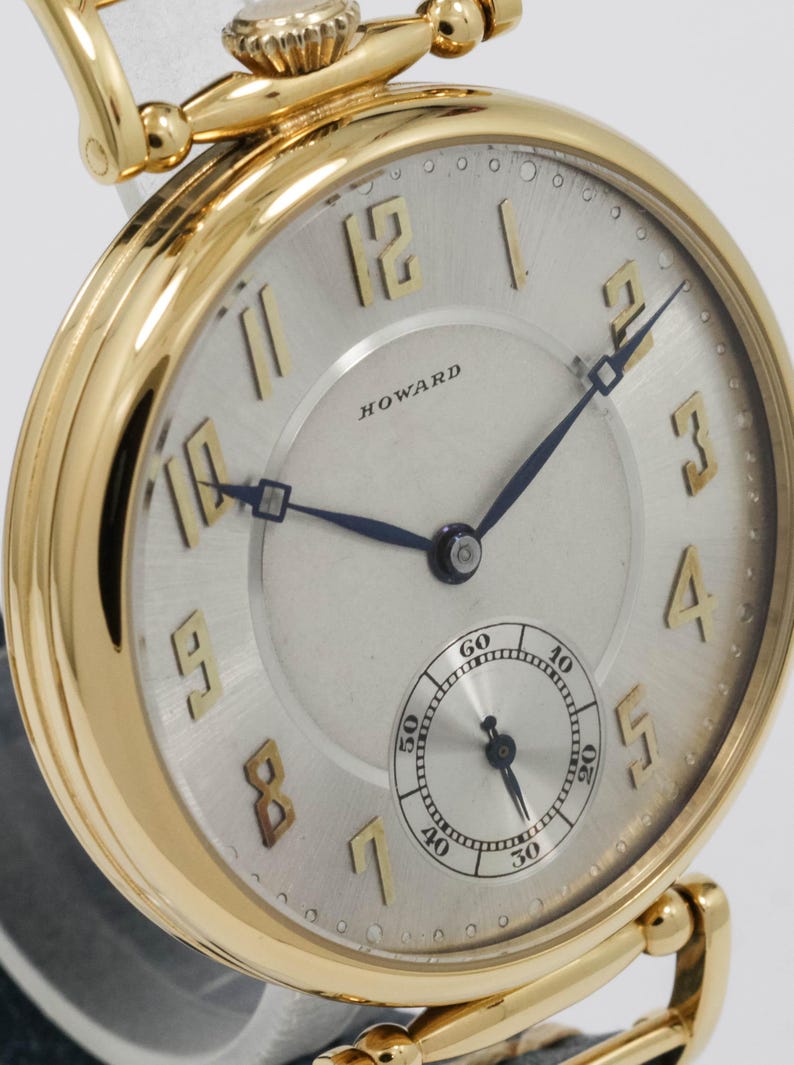 May include: A gold pocket watch with a silver face. The watch has black hands, gold numerals, and a small seconds dial. The word "HOWARD" is printed on the face. The watch has a gold crown and a gold chain attachment.