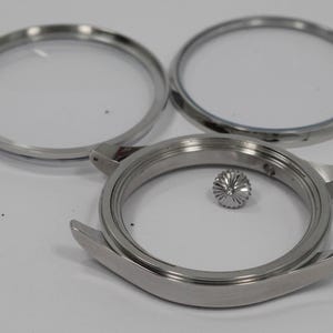 May include: A collection of disassembled watch parts. The image shows a stainless steel watch case, two clear watch crystals, and a crown. The watch case has curved lugs and a circular opening. The crown is a small, textured, silver-coloured piece.