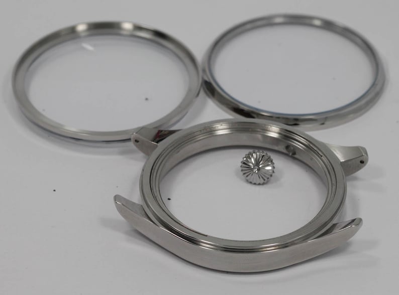 May include: A collection of disassembled watch parts. The image shows a stainless steel watch case, two clear watch crystals, and a crown. The watch case has curved lugs and a circular opening. The crown is a small, textured, silver-coloured piece.
