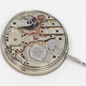 Puede incluir: Vista detallada de un movimiento de reloj antiguo, que muestra intrincados engranajes, joyas de rub&iacute; y una caja de metal plateado. Los componentes mec&aacute;nicos est&aacute;n dispuestos en un patr&oacute;n circular, con una tija de cuerda que se extiende desde el lateral. El mecanismo del reloj est&aacute; sobre un fondo blanco.