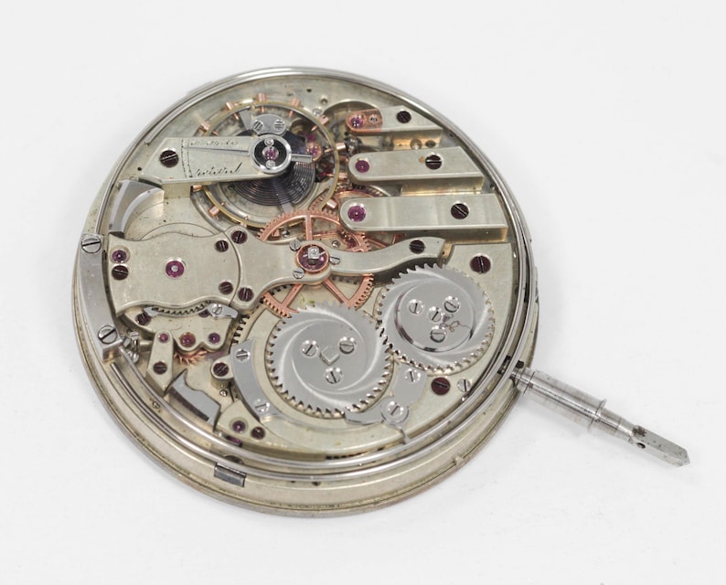Puede incluir: Vista detallada de un movimiento de reloj antiguo, que muestra intrincados engranajes, joyas de rub&iacute; y una caja de metal plateado. Los componentes mec&aacute;nicos est&aacute;n dispuestos en un patr&oacute;n circular, con una tija de cuerda que se extiende desde el lateral. El mecanismo del reloj est&aacute; sobre un fondo blanco.