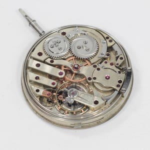 Puede incluir: Un movimiento de reloj plateado con engranajes, resortes y joyas de rub&iacute; expuestos. El intrincado mecanismo presenta m&uacute;ltiples capas de componentes, incluido un v&aacute;stago de cuerda. El reloj est&aacute; sobre un fondo blanco.
